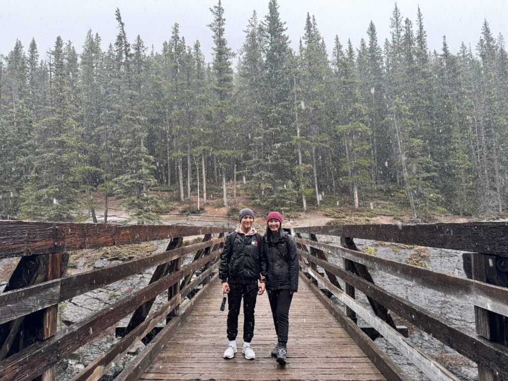 Two people stand on the wooden Stewart Canyon Bridge in Banff National Park, Alberta during snowfall, surrounded by dense evergreen trees. They wear warm clothing, creating a serene, wintry atmosphere.