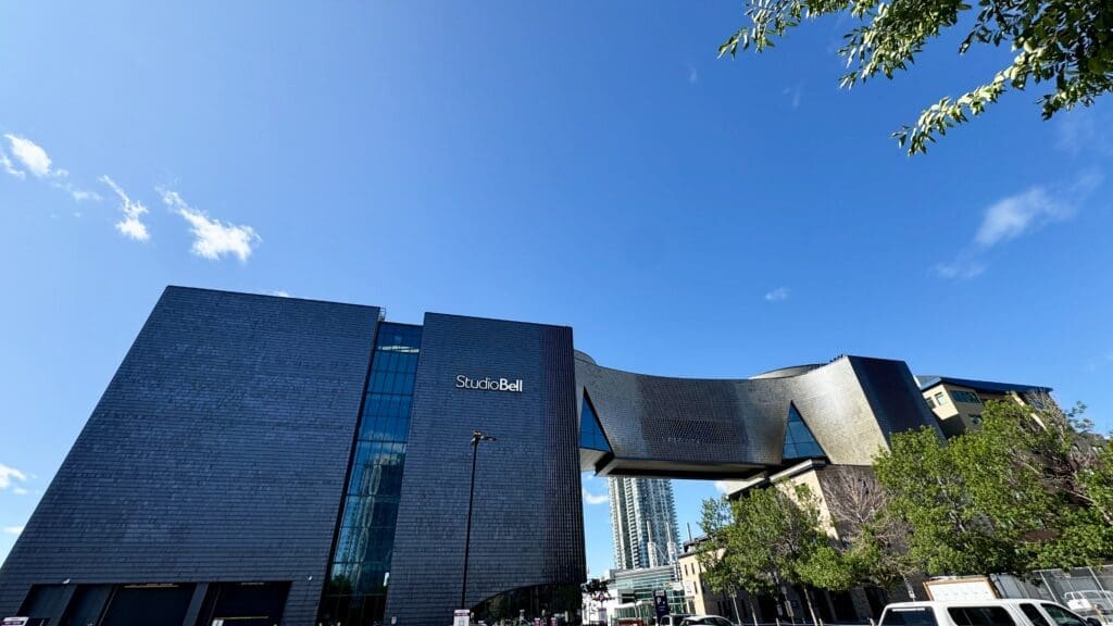 Modern building with sleek black facade labeled "Studio Bell," in Calgary, Alberta set against a clear blue sky. Trees and a few white vehicles in the foreground.