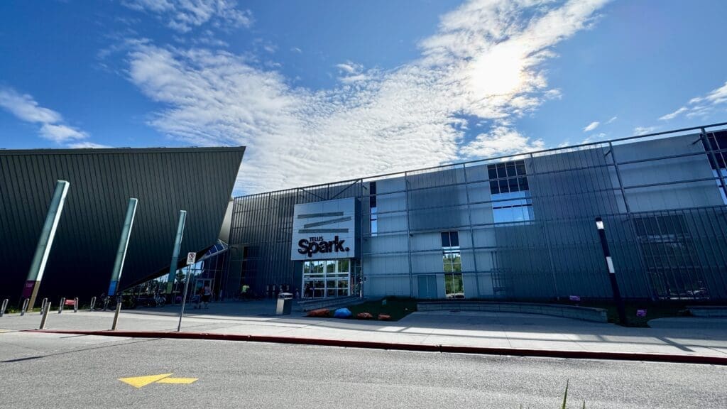 Wide-angled view of modern building in Calgary, Alberta with angular design, labeled "TELUS Spark." Bright blue sky, scattered clouds, and sun create a vibrant atmosphere.