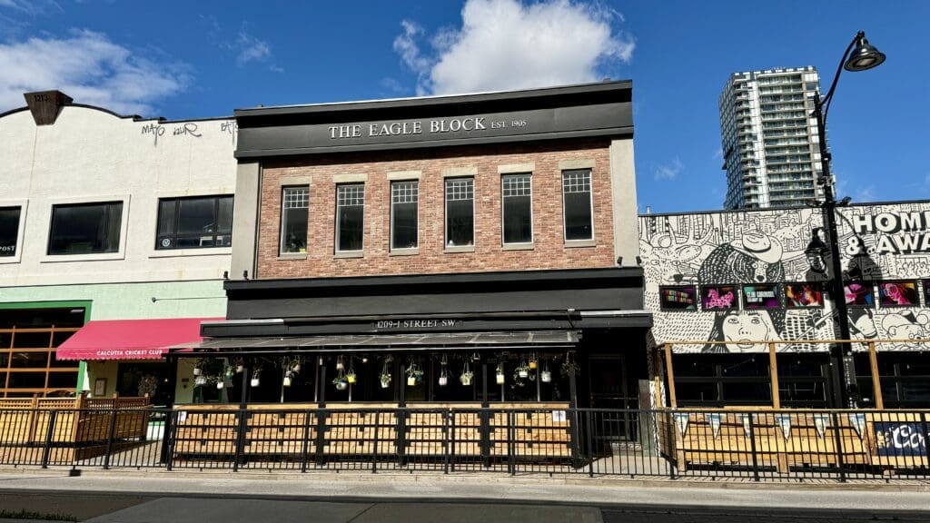 A two-story brick building labeled "The Eagle Block" with the restaurant Ten Foot Henry inside in Calgary, Alberta, beside a mural-covered structure with colorful signage. Bright, sunny day with blue sky.