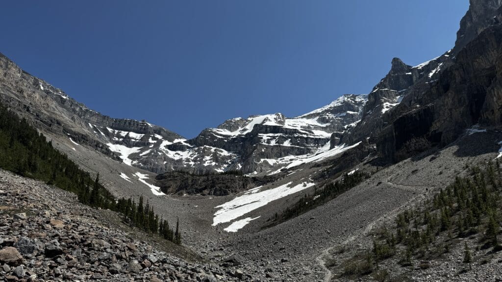 Snow-dusted Stanley Glacier in Kootenay National Park, BC under a clear blue sky, with rugged rocky terrain and scattered evergreen trees in the foreground, evoke a serene, majestic feel.