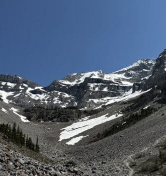 Snow-dusted Stanley Glacier in Kootenay National Park, BC under a clear blue sky, with rugged rocky terrain and scattered evergreen trees in the foreground, evoke a serene, majestic feel.