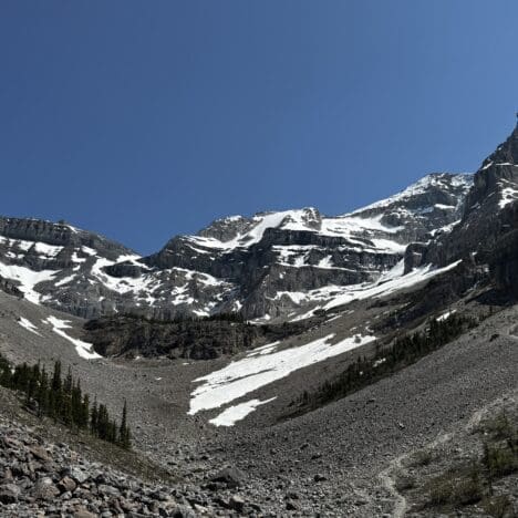 Snow-dusted Stanley Glacier in Kootenay National Park, BC under a clear blue sky, with rugged rocky terrain and scattered evergreen trees in the foreground, evoke a serene, majestic feel.