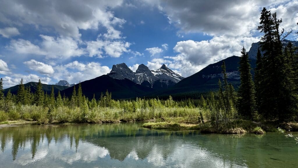 Mountain landscape with Three Sisters in Canmore, Alberta under a partly cloudy sky, reflected in a tranquil lake. Dense green forest lines the shore, conveying serenity.
