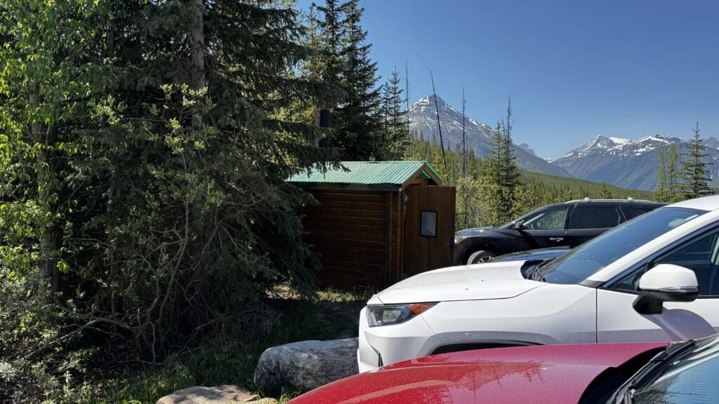 Cars are parked on a sunny day near a small wooden cabin with a green roof at Stanley Glacier Trailhead in Kootenay National Park, BC, surrounded by dense trees. Snow-capped mountains are visible in the background.