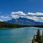 Scenic view of Two Jack Lake in Banff National Park, Alberta surrounded by dense evergreen forests, with majestic Mount Rundle under a clear blue sky and scattered clouds. Peaceful and serene.