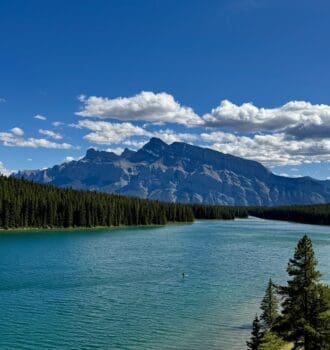 Scenic view of Two Jack Lake in Banff National Park, Alberta surrounded by dense evergreen forests, with majestic Mount Rundle under a clear blue sky and scattered clouds. Peaceful and serene.