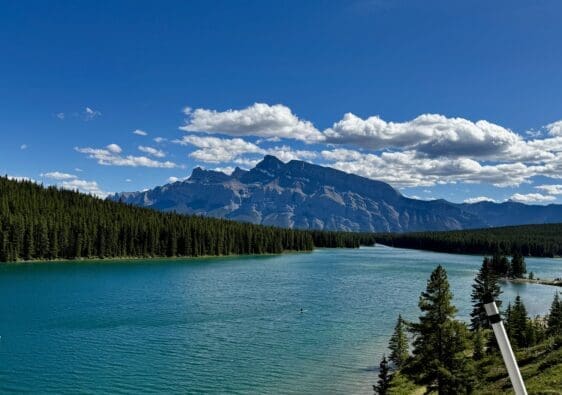 Scenic view of Two Jack Lake in Banff National Park, Alberta surrounded by dense evergreen forests, with majestic Mount Rundle under a clear blue sky and scattered clouds. Peaceful and serene.