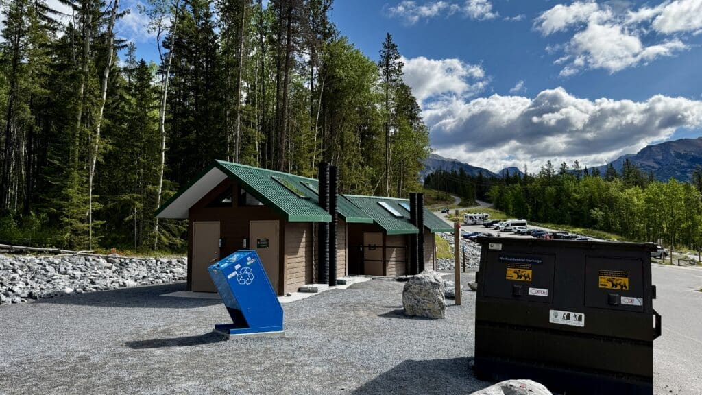 Public restroom and recycling bin with green roofs set against a forest backdrop at Grassi Lakes Trailhead in Kananaskis, Alberta. Blue sky, scattered clouds, parked cars, and mountains in the background.