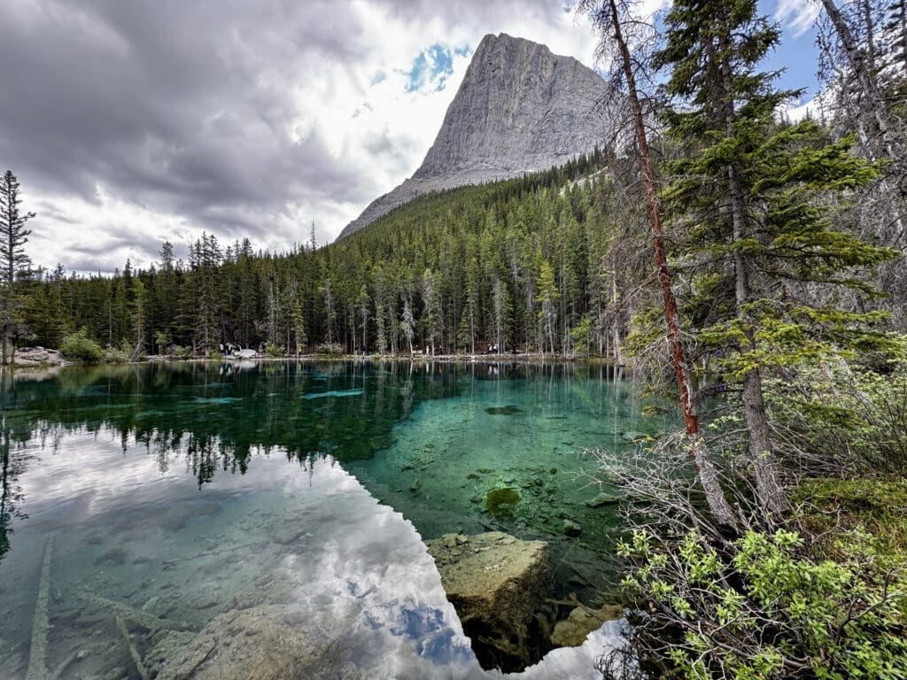 Clear turquoise Grassi Lakes in Kananaskis, Alberta reflecting pine trees and a rocky mountain under a cloudy sky
