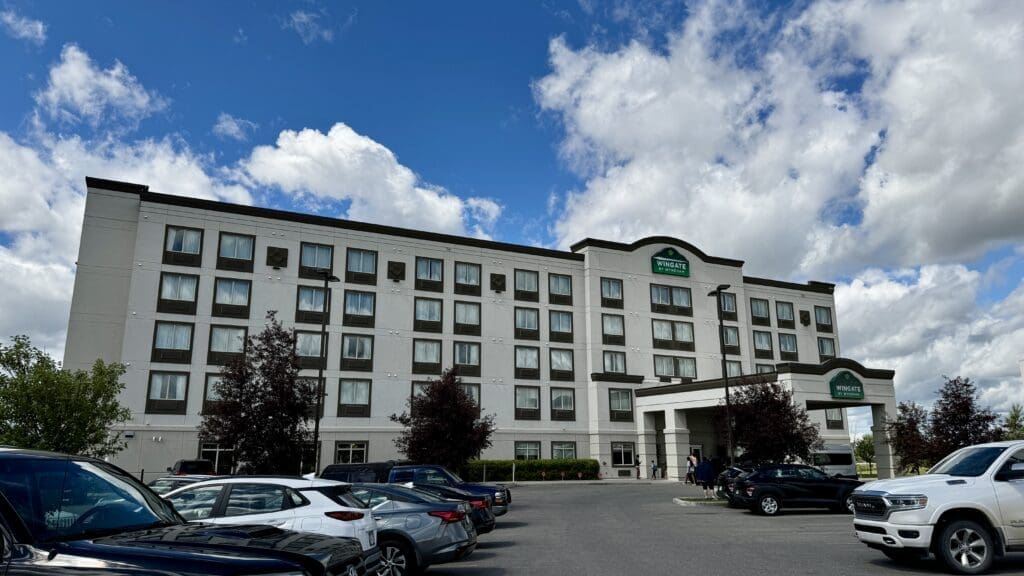 Five-story Wingate by Wyndham in Calgary Airport, Alberta with tan facade and dark window frames under a blue sky with clouds. Cars are parked in the lot, creating a welcoming atmosphere.