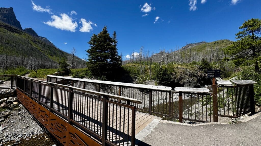 A wooden bridge with metal railings at Red Rock Canyon in Waterton Lakes National Park, Alberta crosses a rocky stream, set against a backdrop of lush green hills and a bright blue sky with fluffy clouds.
