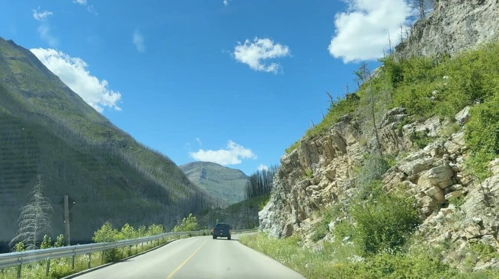 Akamina Parkway in Waterton Lakes National Park, Alberta with a black SUV traveling forward, flanked by lush green slopes and rocky cliffs under a clear blue sky with fluffy clouds.