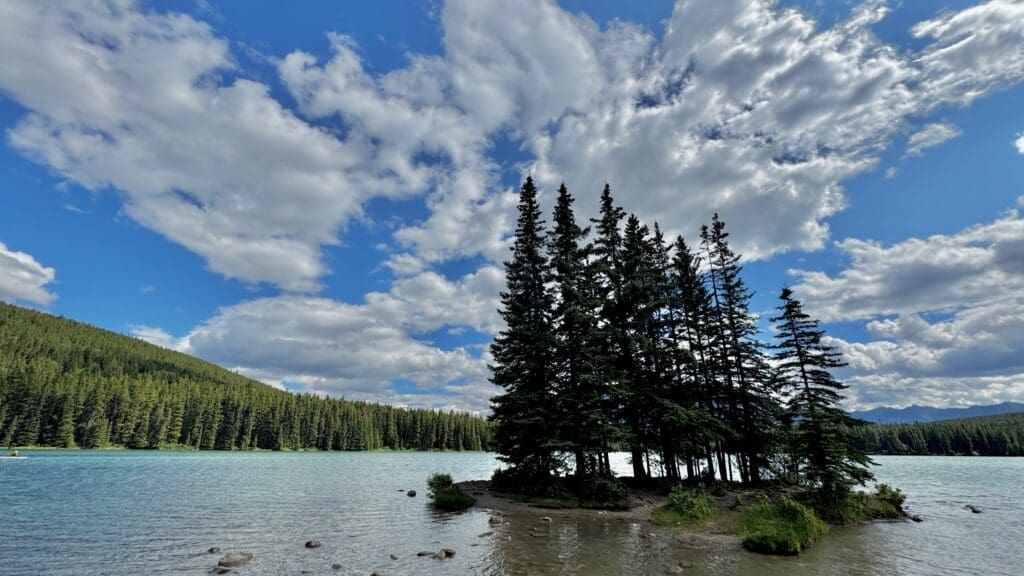 Two Jack Lake in Banff National Park, Alberta with calm, turquoise water surrounded by dense green pine trees. Fluffy white clouds dot the blue sky, conveying a peaceful atmosphere.