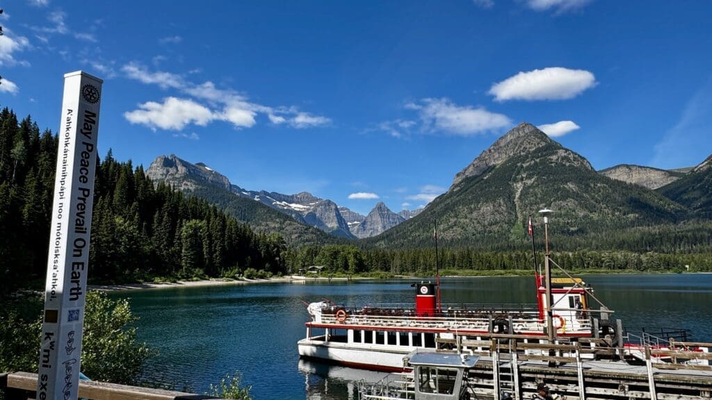 A peaceful mountain lake scene with a "May Peace Prevail On Earth" sign in the foreground. The Waterton Shoreline Cruise is docked, surrounded by lush pine forests and majestic mountains under a clear blue sky.