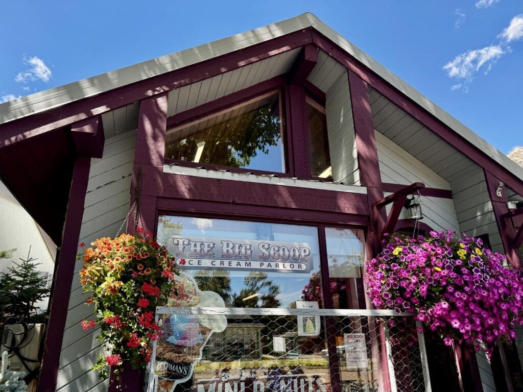 A charming ice cream parlor with a triangular roof and maroon trim in Waterton Townsite, Alberta. Colorful flower baskets hang by the entrance, contrasting with the clear blue sky.