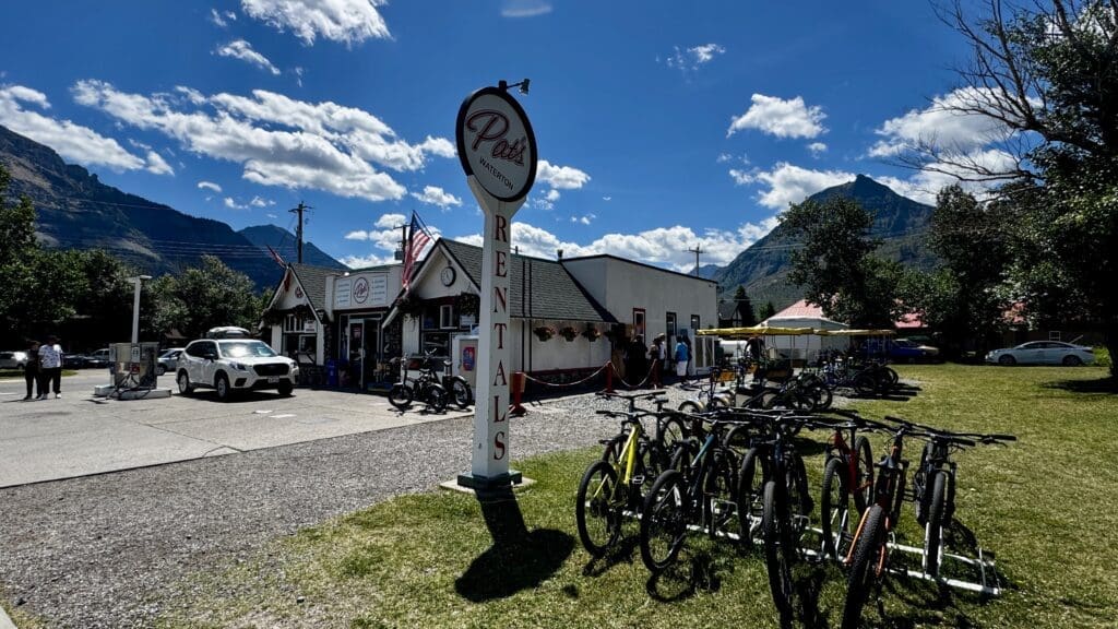 A bike rental shop in Waterton Townsite in Waterton Lakes National Park, Alberta with a "Rentals" sign stands against a backdrop of mountains and a clear blue sky. Bicycles and a car are in the foreground.