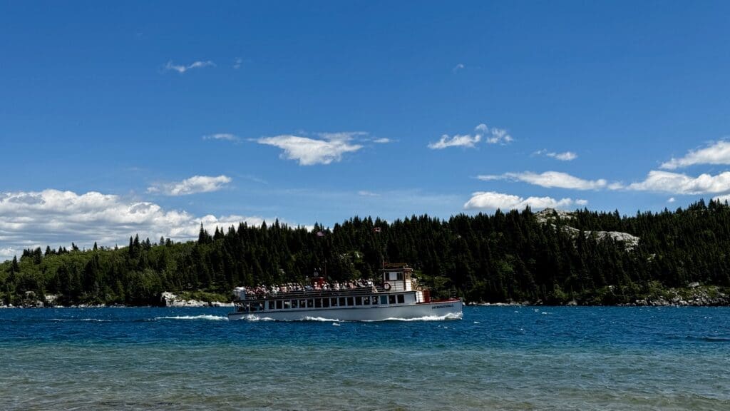 The white Waterton Shoreline Cruise sails on Waterton Lakes in Waterton Lakes National Park, Alberta under a bright sky with scattered clouds. The background features dense green forests and rocky hills.
