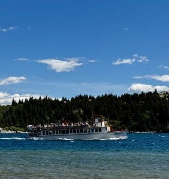 The white Waterton Shoreline Cruise sails on Waterton Lakes in Waterton Lakes National Park, Alberta under a bright sky with scattered clouds. The background features dense green forests and rocky hills.