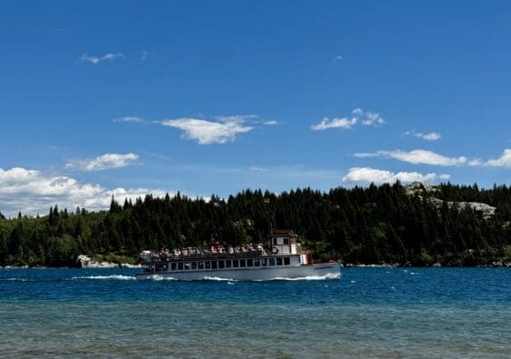 The white Waterton Shoreline Cruise sails on Waterton Lakes in Waterton Lakes National Park, Alberta under a bright sky with scattered clouds. The background features dense green forests and rocky hills.