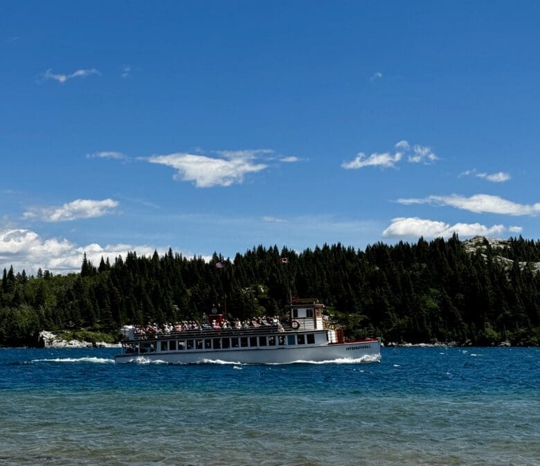 The white Waterton Shoreline Cruise sails on Waterton Lakes in Waterton Lakes National Park, Alberta under a bright sky with scattered clouds. The background features dense green forests and rocky hills.