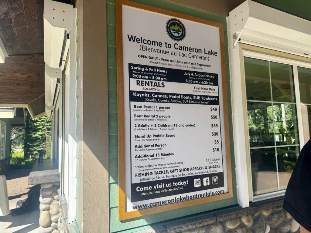 Sign for Cameron Lake Boat Rentals in Waterton Lakes National Park, Alberta offering kayaks, canoes, and more. Prices listed for boat and SUP rentals. Bright day, wooden building.