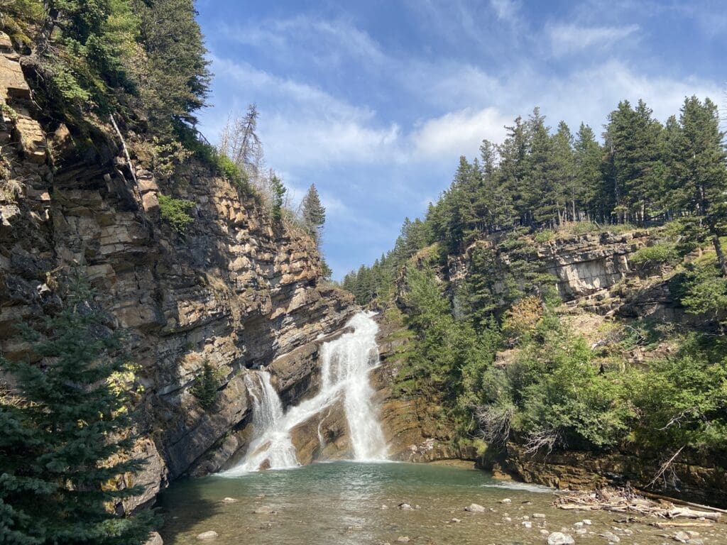 The serene Cameron Falls in Waterton Lakes National Park, Alberta cascades down rocky cliffs into a tranquil pool, surrounded by lush green pine trees under a bright blue sky, conveying peace.