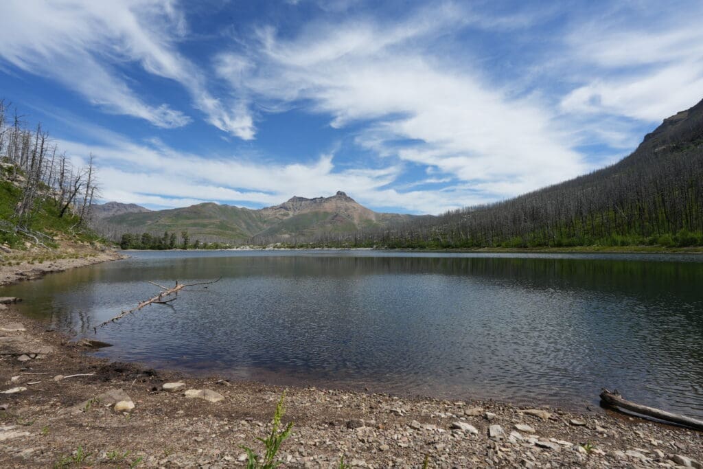 Crandell Lake in Waterton Lakes National Park, Alberta with clear blue sky and wispy clouds. The calm water reflects surrounding green hills and burned trees, creating a tranquil scene.