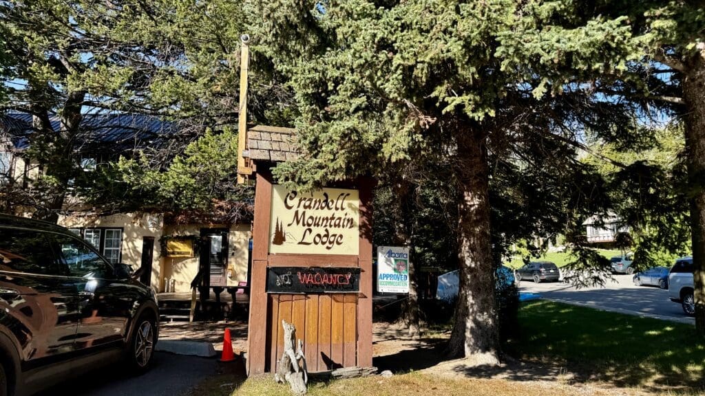 Sign for Crandell Mountain Lodge in Waterton Townsite, Alberta among trees, displaying a "No Vacancy" notice. A parked car is near the entrance, under dappled sunlight.