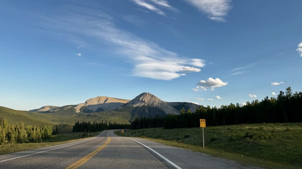 Open road on the way to Forgetmenot Pond in Kananaskis, Alberta leading to distant mountains under a clear blue sky with scattered clouds, surrounded by lush green trees and a speed limit sign on the right.