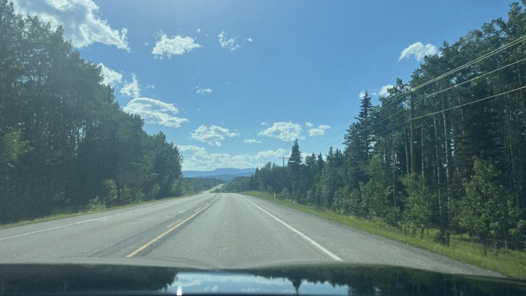 Wide open road heading to Fullerton Loop in Alberta flanked by dense green forests under a clear blue sky with scattered clouds. Sunlight creates a serene and inviting atmosphere.