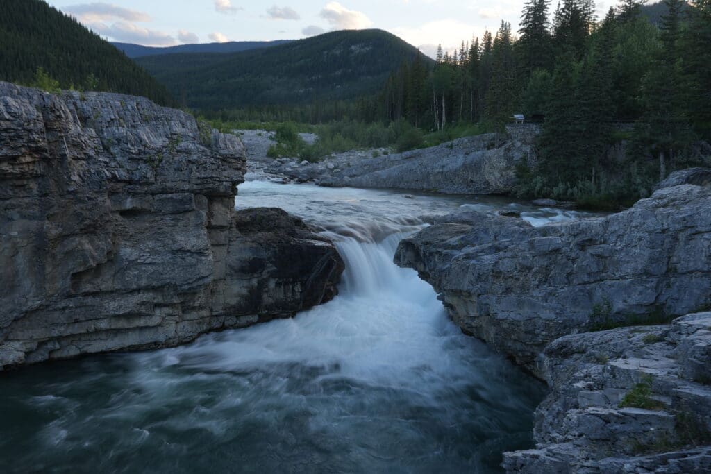Rocky waterfall in Elbow Falls, Alberta scene with rushing water cascading between large gray boulders. Lush pine forest and distant mountains under a cloudy sky at dusk.