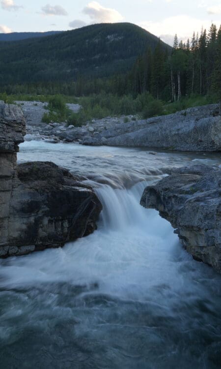 Rocky waterfall in Elbow Falls, Alberta scene with rushing water cascading between large gray boulders. Lush pine forest and distant mountains under a cloudy sky at dusk.