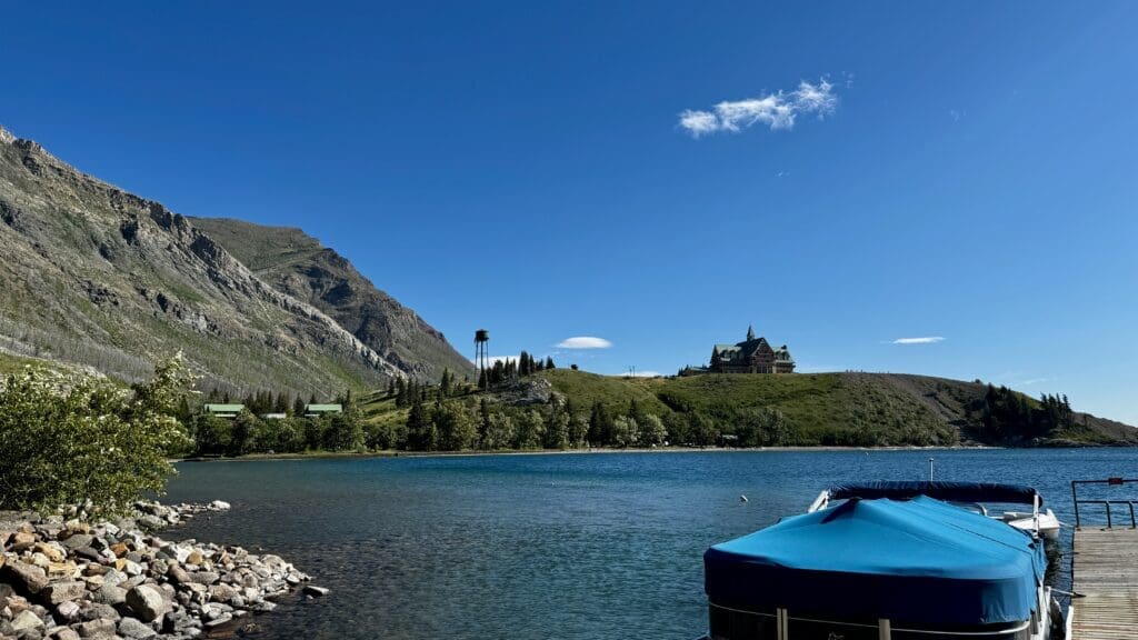 Clear blue lake in Waterton Lakes National Park, Alberta with a rocky shore, boats covered with a blue tarp, green hills, distant rugged mountains, and a castle-like building under a bright, clear sky.