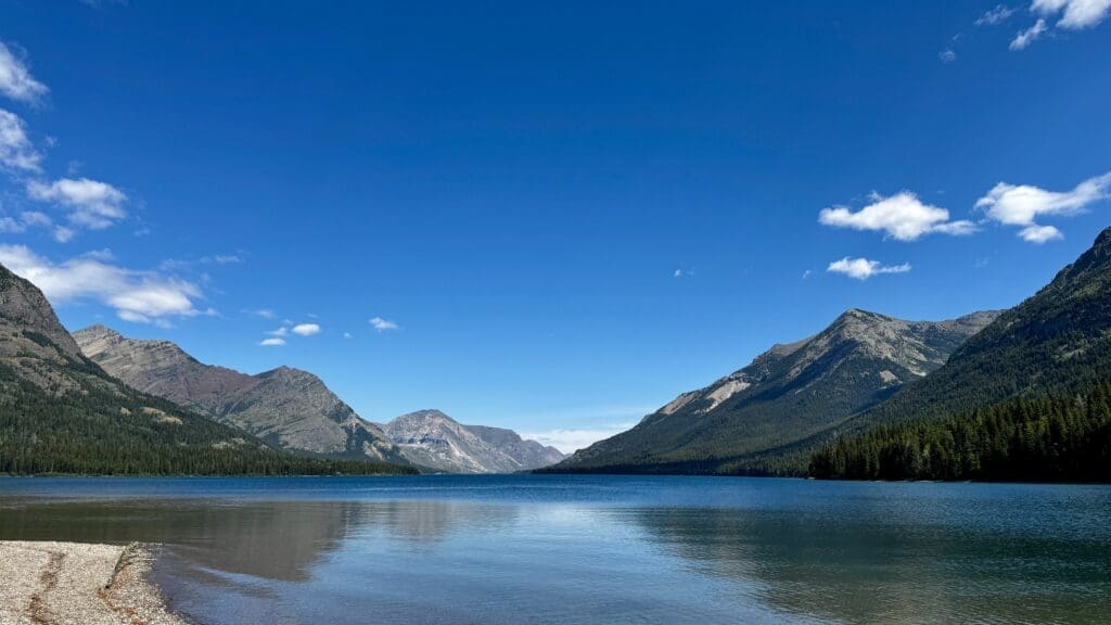A serene mountain lake scene with clear blue water in Goat Haunt in Glacier National Park, Montana, flanked by forested mountains under a bright blue sky with scattered white clouds. Peaceful and natural.