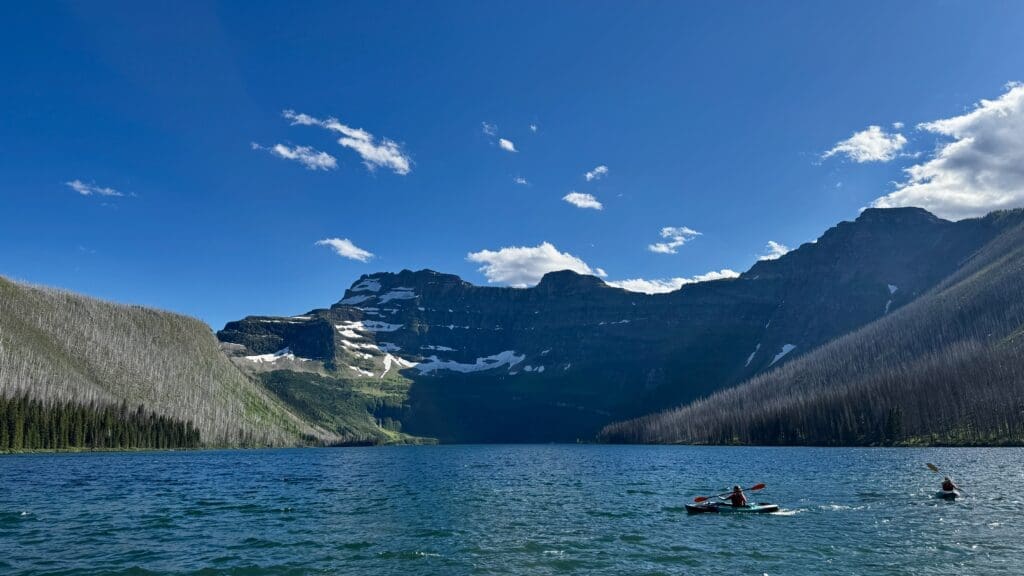 Kayakers paddle on the serene Cameron Lake in Waterton Lakes National Park, Alberta, surrounded by lush green and rugged mountains under a bright blue sky with scattered clouds.