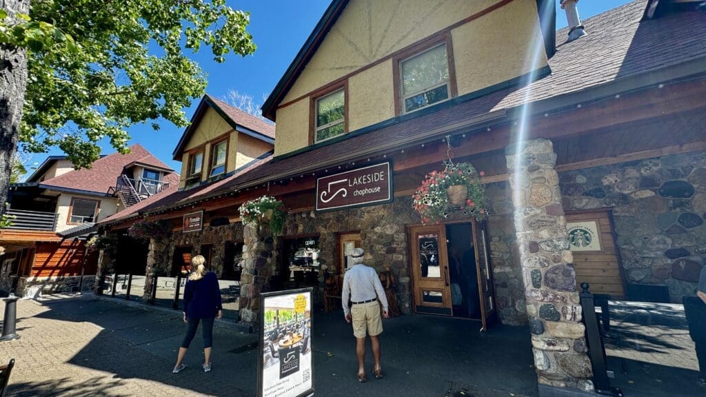 Charming stone building with "Lakeside Chophouse" sign in Waterton Townsite, Alberta, colorful hanging flowers. Two people standing outside under a sunny blue sky. Inviting, rustic atmosphere.