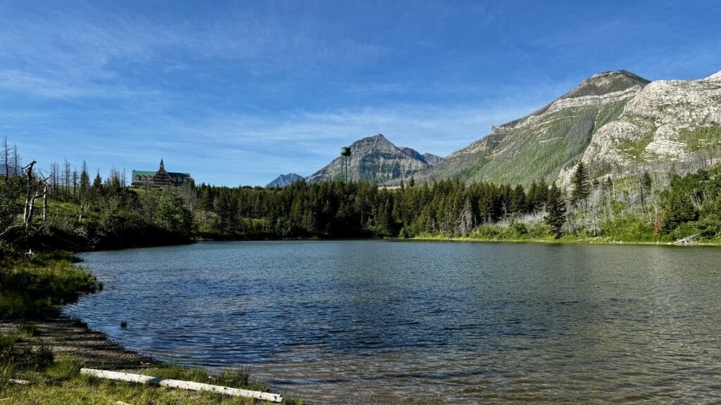 Linnet Lake in Waterton Lakes National Park, Alberta bordered by evergreen trees, with a distant lodge and a water tower against a backdrop of rugged mountains under a clear blue sky.