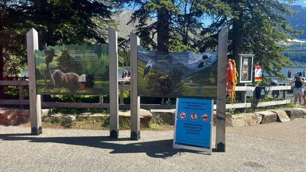 Wooden signs with nature illustrations and information about grizzlies and birds stand by a lakeside path. A blue sign prohibits launching watercraft in Cameron Lake in Waterton Lakes National Park, Alberta.
