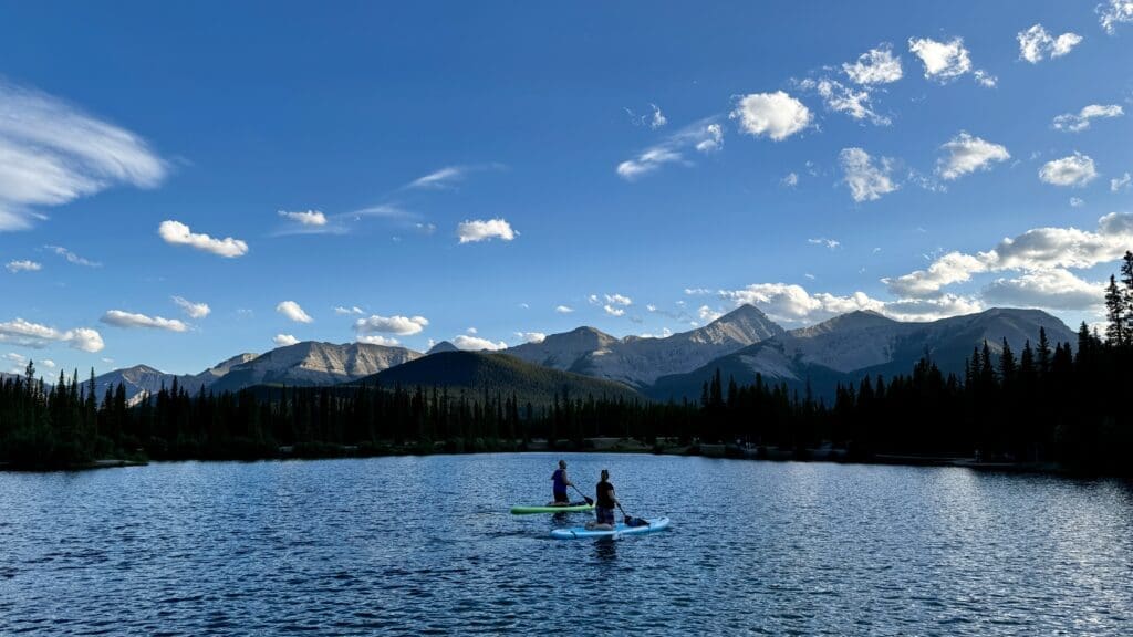 Two people paddleboard on a Forgetmenot Pond in Kananaskis, Alberta under a clear blue sky, surrounded by majestic mountains and pine trees, evoking a sense of tranquility.