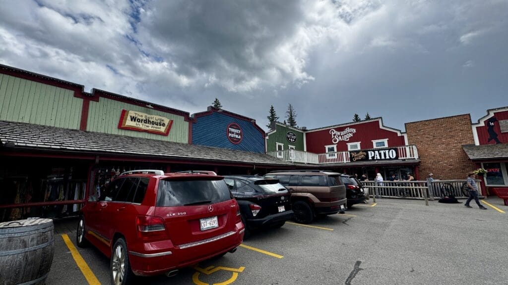 A row of rustic, Western-themed shops in Bragg Creek Hamlet, Alberta with colorful facades and signs, parked cars in front, under a cloudy sky. People gather near a saloon patio.