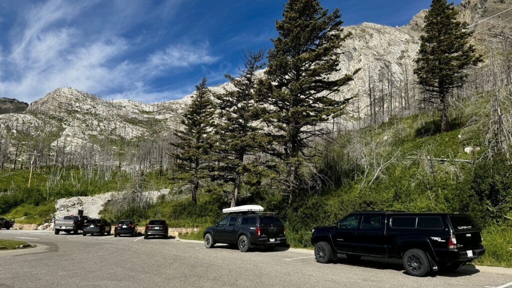Cars parked by a road at Bear's Hump Trailhead in Waterton Lakes National Park, Alberta under a clear blue sky with scattered clouds. Rugged mountains and tall evergreen trees create a peaceful, natural setting.
