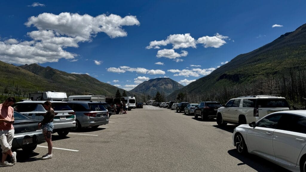Parking lot at Red Rock Canyon in Waterton Lakes National Park, Alberta with cars on both sides, surrounded by green mountains under a bright blue sky with fluffy clouds. Two people stand talking on the left.