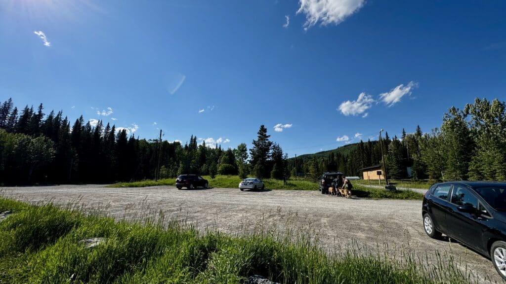 A serene parking area of Fullerton Loop Trailhead in Kananaskis, Alberta bordered by lush pine trees with a clear blue sky. Several parked cars, people with dogs, and a wooden cabin create a peaceful, sunny day.