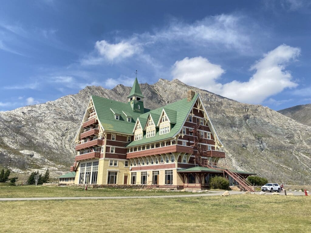 The large, historic Prince of Wales Hotel in Waterton Lakes National Park, Alberta with a steep green roof stands against a rocky mountain backdrop under a bright blue sky with scattered clouds, conveying a serene, majestic atmosphere.
