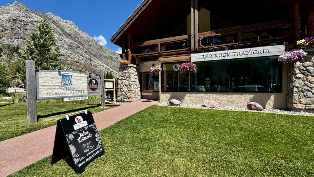 Charming wooden restaurant Red Rock Trattoria in Waterton Townsite, Alberta with stone facade, hanging flowers, and outdoor signs. Set against a mountain backdrop under a clear blue sky.