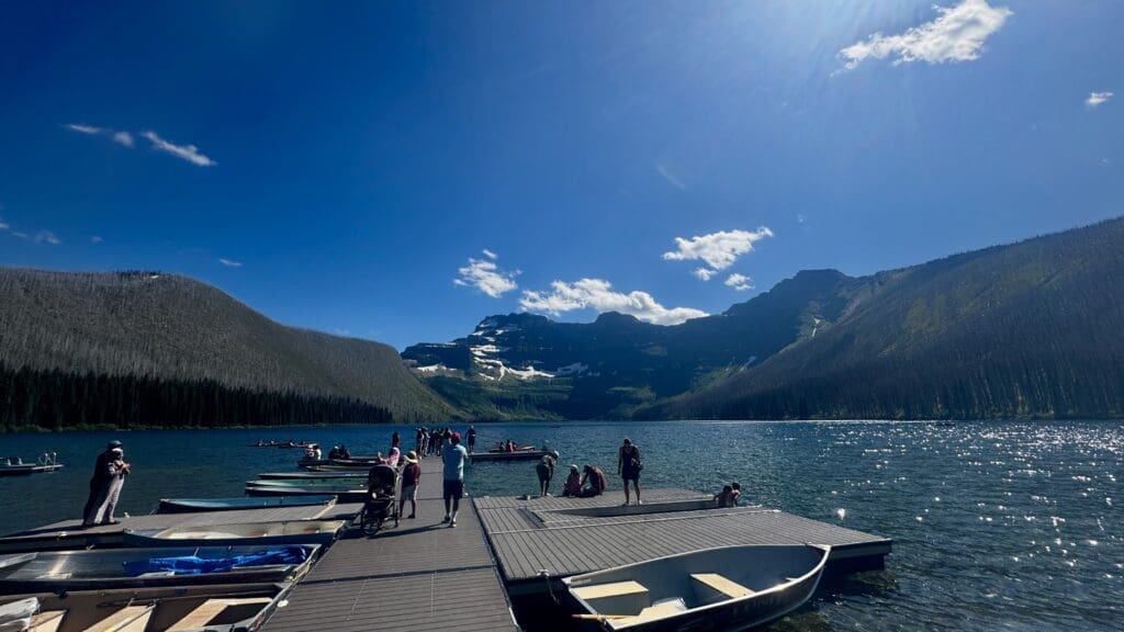 Boat dock on Cameron Lake in Waterton Lakes National Park, Alberta with people gathered, surrounded by majestic mountains under a bright blue sky. A serene and lively atmosphere.