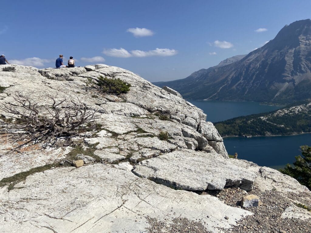 Two people sit on a rugged, rocky cliff overlooking a vast lake in Waterton Lakes National Park, Alberta and steep mountains under a clear blue sky, evoking tranquility and awe.