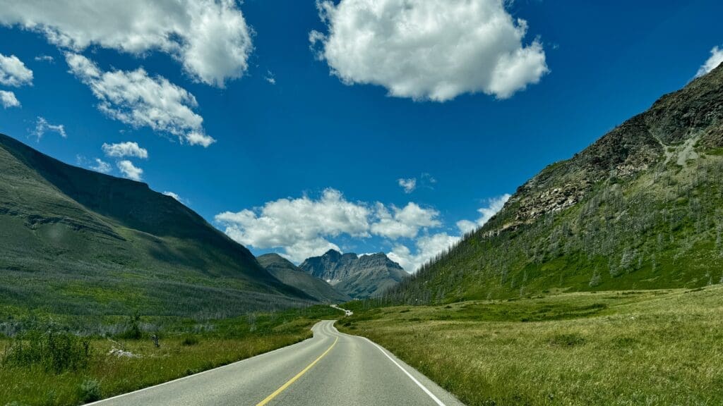 The Red Rock Parkway in Waterton Lakes National Park, Alberta leads through a lush green valley flanked by rugged mountains under a bright blue sky with fluffy white clouds, evoking tranquility.