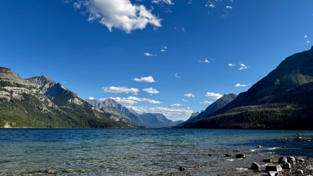 Waterton Lake, Alberta with clear, rippling water reflects towering mountains under a bright blue sky with scattered clouds. A person sits on the rocky shore.